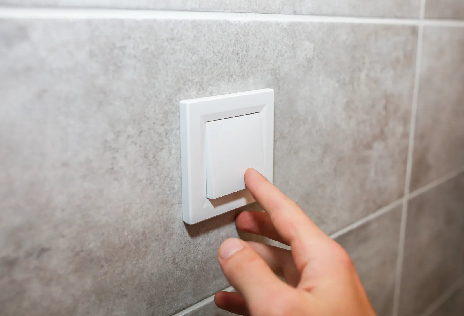 Hand pressing a white light switch on a tiled bathroom wall